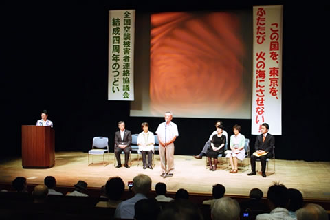 集会の舞台全景。「この国を、東京を、ふたたび 火の海にさせない」「全国空襲被害者連絡協議会結成四周年のつどい」の垂れ幕が下がっている。舞台上には数人分の椅子が並べられており、立って話している一人以外は着席している。