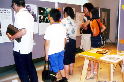 武蔵野市役所ロビーでの展示。ついたてに原爆展パネルが掲示されており、通路を挟んで置かれた小さいテーブルの上にも資料がある。原爆展パネルを閲覧する人たちがついたての前をうめるように列を作っている。