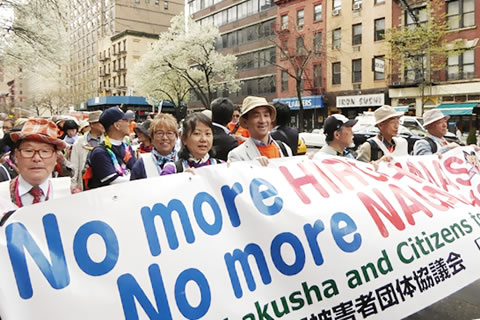ニューヨーク市街を横に8人並ぶ列を作って行進する、日本被団協と生協連の代表団。先頭には「No more HIROSIMAS No more NAGASAKIS」と書かれた、縦1メートル、横5メートルほどの横断幕を掲げている。
