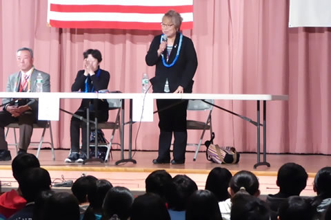 講堂のアメリカ国旗と日の丸が掲げられた舞台上から、日本人学校の生徒たちに語る東友会メンバー。