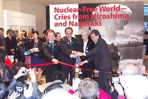 「Nuclear-Free World― Cries from Hirosima and Nagasaki(核のない世界を― 広島・長崎からの叫び)」と書かれた、縦横3メートルほどの看板の前で行われたテープカット。大きいはさみを、被爆者代表などが持ち、赤いテープを切る場面。大看板には、キノコ雲の写真がいっぱいに使われている。