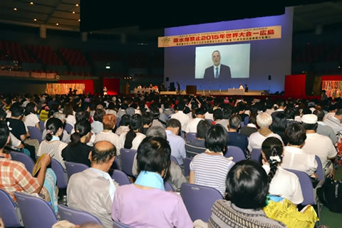 広島の全体集会。体育館に並べられた座席を参加者が埋めている。舞台上のスクリーンには話をする人が投影されている。