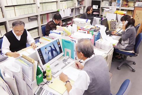 書類の収められたケースでいっぱいの棚を背景に、パソコンや書類が乗った事務机で仕事をする職員たち