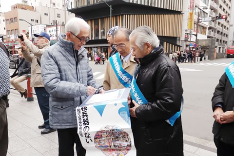「核兵器のない平和で公正な世界を」と書かれたポスターを手持ちの署名板の前に垂らし署名を呼びかける被爆者と、ペンを持ちながら署名用紙を読む人