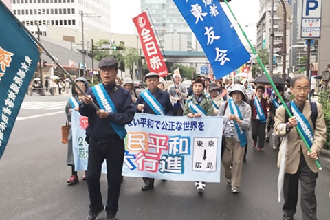 「われら生命もて ここに証す 原爆許すまじ」の旗、「一般社団法人 東友会」を掲げ、「国民平和大行進」の先頭に立つ東京の被爆者たち。「平和大行進」の横幕も東友会のたすきを掛けた被爆者5人が持っている。後ろに行進参加の各団体の人びとが連なっている。