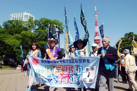 「国民平和大行進」の横幕を、東友会のたすきをかけた被爆者と通し行進者が持って歩いている。後ろには参加団体の旗やのぼりがいくつも立っており、背景に東京タワー。