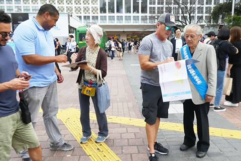 「ハチ公前広場」で、署名用紙を載せた板を持ち、たすきを掛けるなどして署名を呼びかける人たち。背の高い外国人が足を止め、署名している。