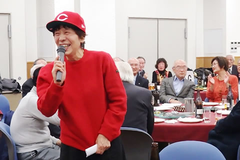 広島カープ球団の帽子をかぶり、自席で立ちマイクを持って笑顔で報告する地区の会の方