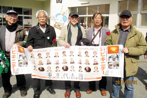 3・1ビキニデー集会のポスター、被爆者国際署名のポスターを横断幕のようにつないだものをみんなで持つ、5人の東京代表団