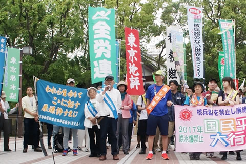たすきを掛けた被爆者が挨拶をしている場面。広島まで歩く「通し行進者」や横断幕を持つ人たち、東友会の旗、参加した各団体ののぼりが写っている。