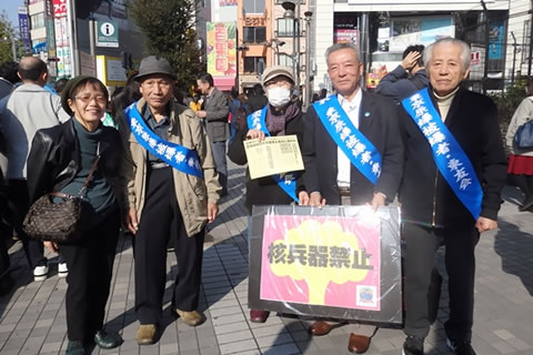 「東友会」のたすきを掛けてた被爆者と、一緒に行動した参加者が笑顔で並んだ写真。一人の被爆者は「核兵器禁止」と大きく書かれた鮮やかなポスターを持っている