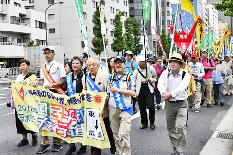 車道を進む行進の先頭で横断幕を持っているのは、たすきを掛けた被爆者と、広島まで歩く「通し行進者」などのひとたち。後ろに続く長い列からはさまざまな団体の登りがかかげられている。