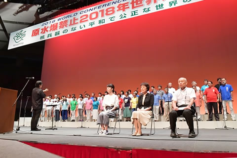 原水爆禁止2018年世界大会、閉会総会の舞台。椅子に座った発言者の後ろに合唱隊が並び、指揮者に合わせ演奏中。