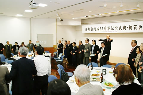 東友会結成55周年記念式典・祝賀会と書かれた看板の掲げられた会場、立って腕を組み合唱する参加者。