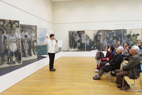 絵画が展示された会場で、椅子に座って説明を聞く参加者
