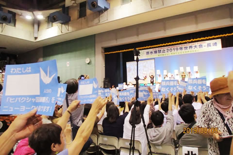 原水爆禁止2019年世界大会、閉会総会の舞台。「核兵器をなくそう」と書かれた紙を、舞台上に立つ人たちと会場の椅子に座った参加者がみんな掲げている。