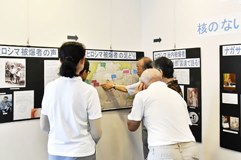 原爆投下当時の地図など展示物が壁に掲示されている会場。数人の参観者が説明を聞いている。