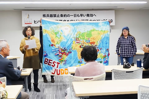 世界地図の描かれた布を広げて参加者に示しながら活動報告する参加団体。布は縦2メートル近く、横は2メートルほど。地図上、核兵器禁止条約を批准した国の上に国旗と国名が描かれている