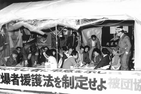 「被爆者援護法を制定せよ 被団協」などと書かれた横断幕を前に、テントの中で座り込みを行う被爆者たち。
