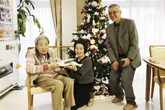 椅子に座る訪問先の方にお見舞い金と編み物を渡す訪問者ら。背景にクリスマスツリー。