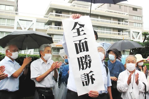 「全面勝訴」と大きく縦書きされた紙を両手で掲げる弁護士と、その周囲で拍手する人たち。雨が降っており、傘をさしている。