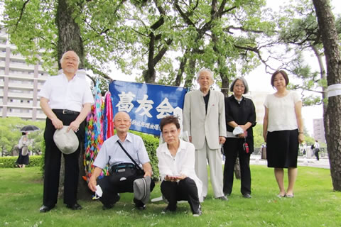 広島の平和公園内、クスノキの周りに集まった葛友会代表と東友会代表。千羽鶴と葛友会の旗がクスノキのそばに置かれている。