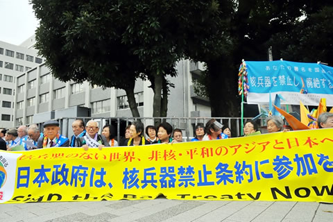 「日本政府核は、兵器禁止条約に参加を」など書かれた横断幕を広げ歩道にに座る被爆者ら。千羽鶴や、人の肩幅よりも大きい折り鶴なども掲げられている。