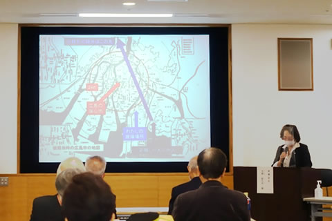 部屋前方の大きいスクリーンには爆心地などが示された広島市の地図が投影されている。被爆者はその脇で座ってマイクを使って証言している。