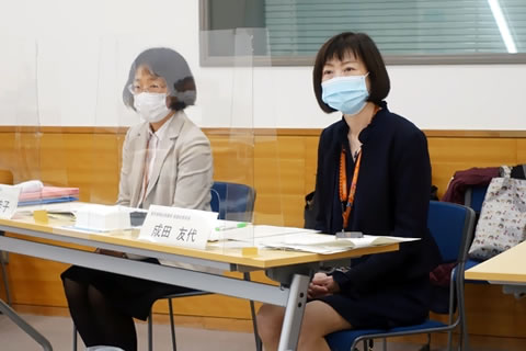 間に透明なついたての立てられた長机に着席する成田部長と堂園課長。二人とも使い捨てマスクを着けている。