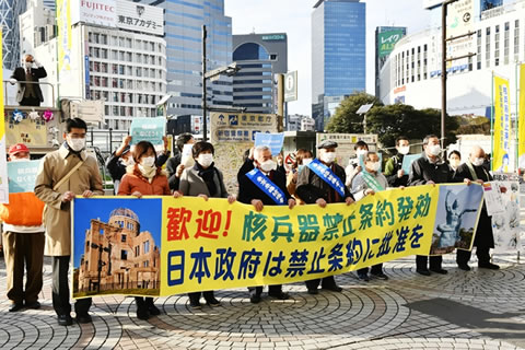 「歓迎！ 核兵器禁止条約発効 日本政府は禁止条約に批准を」(原文ママ)と書かれた横断幕を並んで掲げ持つ人たち、その後ろや周囲で紙を掲げる人たち、宣伝カーの上で訴える人たちなどが写っている。横断幕を持つ人のうち被爆者はたすきを掛けている。