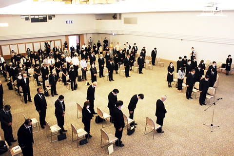 会場に並べられた椅子の前に立ち、黙祷する参加者たち。