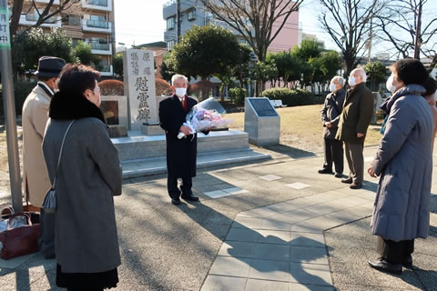 慰霊碑の手前に立つ、東京都原爆被害者協議会の役員ら。1人は献花する花束を抱えて慰霊碑の正面に立ち、慰霊碑を囲んで半円形に並んで立つ他の役員と向き合っている。