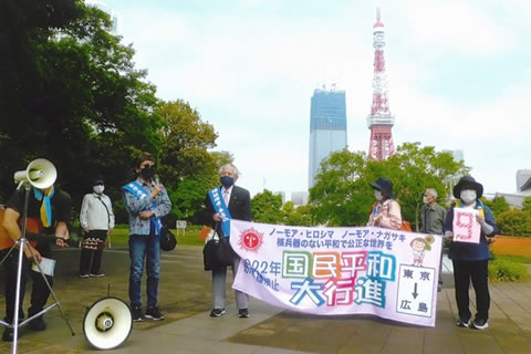 「国民平和大行進」と書かれた横断幕を、2人が両端を持って広げている。うち1人はたすきを掛けた被爆者。その横に立って、別のたすきを掛けた被爆者がハンドマイクを使って話をしている。その他数人が周囲に立っている。背景には東京タワーとビルが写っている。
