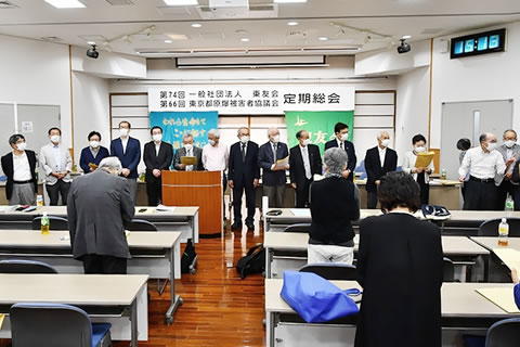 会場前方、「第74回 一般社団法人東友会 第66回 東京都原爆被害者協議会 定期総会」と大きく横書きされた看板が天井近くに下げられている。その手前に、新役員らが横一列に並んで席の方を向いて立っている。並べられた長机の席で出席者も起立している。