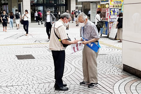 東友会のたすきを掛けた被爆者が街頭に立って、「日本政府に核兵器禁止条約の署名・批准を求める署名」と書かれた紙を取り付けた署名版を持っている。その前に立ってペンをとり、署名をしている人がいる。