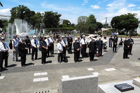 炎天下、石のタイルの敷き詰められた広場に並んで立つ参列者たちを慰霊碑側から撮った写真。。背後には噴水がある。