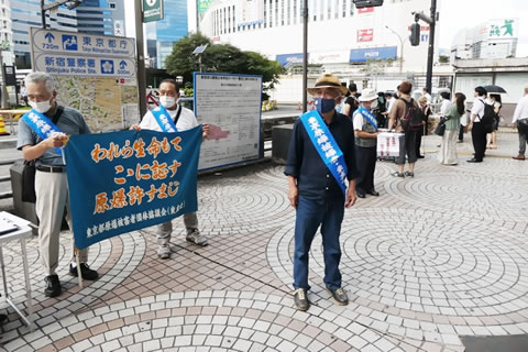 東友会のたすきを掛けた被爆者が街頭に立っている。うち2人が「われら生命もて ここに証す 原爆許すまじ」と書かれた東友会の旗の両端を持ち、体の前に広げている。「日本政府に核兵器禁止条約の署名・批准を求める署名」と書かれた紙を取り付けた署名版を持っている被爆者もいる。