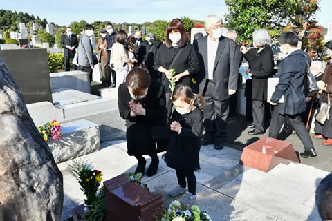 「原爆被害者の墓」の周囲参列者たちが集まっている。うち一人の大人と一人の子どもが、墓の前に立って手を合わせている。手を合わせている大人は、子どもの目線に合わせるように腰をかがめている。