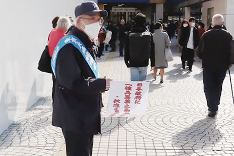 たすきをかけ、「日本政府に核兵器禁止条約の署名・批准を求める署名」と書かれた紙を取り付けた署名版を持っている被爆者が、通行人に呼びかけている。
