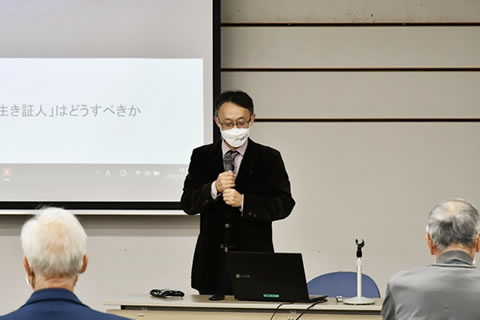 立ってマイクを使いながら話す山田講師。背後のスクリーンが一部写っている。