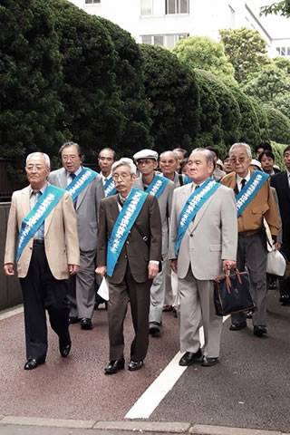 「東京都原爆被害者団体協議会（東友会）」と書かれたたすきをかけ、4列ほどに並んで歩道を歩く原告たち。先頭は4人でなく3人で、原告団長1名と原告副団長2名。