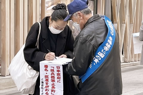 たすきをかけて署名板を持つ被爆者が、ペンを持って署名する通行人と会話している。