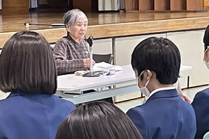 長机に着席して話をする神戸さん。机のかげになっていて、座っているのが車いすかどうかは写真ではわからない。写真手前には、椅子に座った生徒たちの背中が写っている。