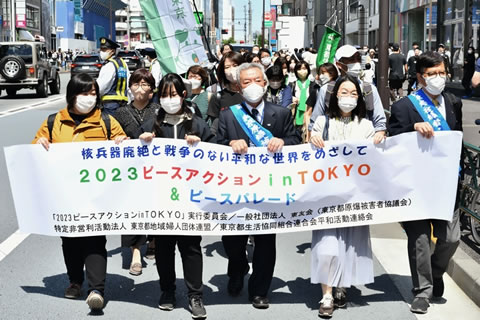 
 「2023 ピースアクション in TOKYO & ピースパレード」など横書きれた横断幕を先頭に街を歩く集会参加者たち。横断幕を持つ5人のうち2人がたすきをかけた被爆者。後ろには、団体名の書かれたのぼりなどを持った人を含め、列が続いている。