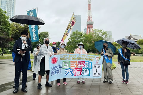 「国民平和大行進」と大きく書かれた横断幕を、4人が持って広げている。うち1人はたすきを掛けた被爆者。横断幕の横に立ってマイクを使って話をしている人やその他周囲に立つ人は、傘をさしたり雨合羽を着たりしている。更新に参加している団体ののぼりも見える。遠景に東京タワーとビルが写っている。