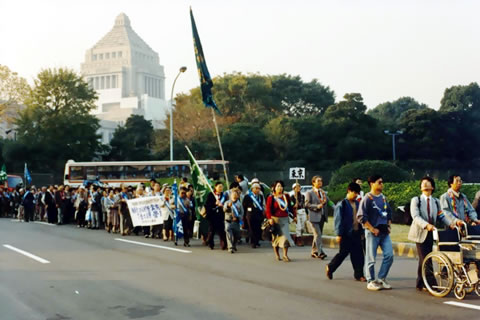 たすきを掛け、旗を持つなどして歩く被爆者たち。背景に国会議事堂が写っている。
