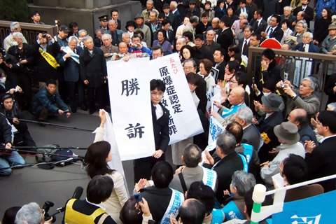 東京地裁前を埋める原告・弁護団・支援者とマスコミの取材陣。多数のカメラとマイクが原告に向けられている。弁護士は「勝訴」の文字などを掲げている。
