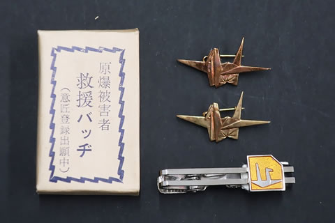 「原爆被害者 救援バッヂ （意匠登録出願中）」と書かれたマッチ箱サイズと思われる紙の箱、折り鶴をかたどった金属製のバッジ2つ、図案化された折り鶴の浮き彫りが配されたネクタイピンが写っている写真。