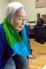 室内で座る、届けられた編み物を首にかけた訪問先の方。