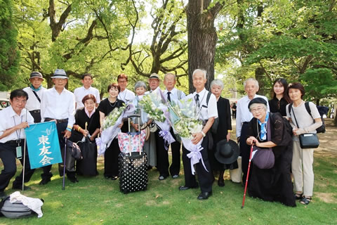 広島平和公園内、木陰での集合写真。20人弱が立って並んでおり、うち数名が花束を抱えている。東友会と書かれた小さい旗を広げて持っている人もいる。